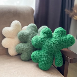 May include: Three plush pillows shaped like shamrocks. The pillows are green and white. The green shamrock pillow is in the foreground and the white shamrock pillow is in the background.