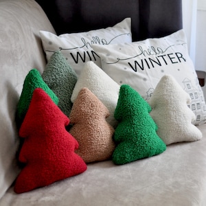 May include: A collection of tree-shaped throw pillows in red, green, sage, tan, and cream colors with a textured sherpa-like finish. Behind them are two white square pillows featuring the black text "hello WINTER" and a line drawing of a house.