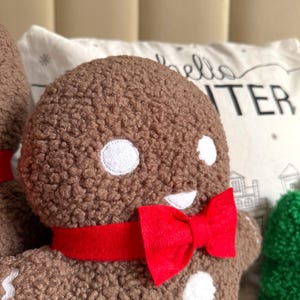 May include: A brown textured plush gingerbread person figure featuring white circular eyes, a white mouth, and a bright red fabric bow tie around the neck. The figure has a soft, fuzzy surface and is positioned in front of a white pillow with text.