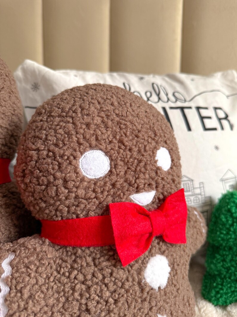 May include: A brown textured plush gingerbread person figure featuring white circular eyes, a white mouth, and a bright red fabric bow tie around the neck. The figure has a soft, fuzzy surface and is positioned in front of a white pillow with text.
