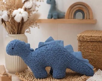 May include: A blue dinosaur plush toy with a textured, curly fabric body and smooth fabric triangular spikes along its back. The dinosaur has a small black eye and a rounded head, standing in a horizontal position with a long, tapering tail.