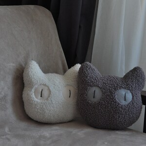 May include: Two plush cat-shaped pillows made of textured sherpa fabric. One pillow is white and the other is dark gray. Both pillows feature large, round, light-colored eyes with vertical black pupils stitched onto the center of the face.