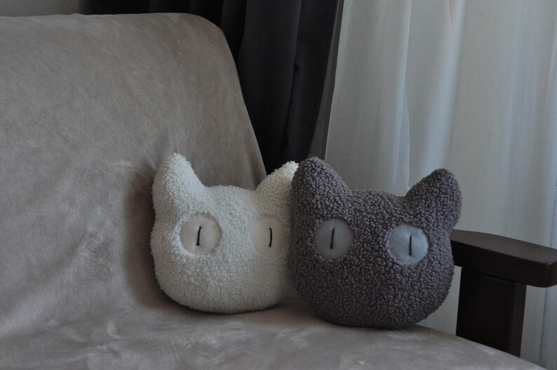 May include: Two plush cat-shaped pillows made of textured sherpa fabric. One pillow is white and the other is dark gray. Both pillows feature large, round, light-colored eyes with vertical black pupils stitched onto the center of the face.