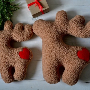 Puede incluir: Dos cojines de peluche marrones con forma de reno, hechos de tela sherpa texturizada. Cada reno presenta un parche de coraz&oacute;n de fieltro rojo brillante en el costado. Los cojines tienen astas distintivas y cuatro patas, con un dise&ntilde;o relleno.