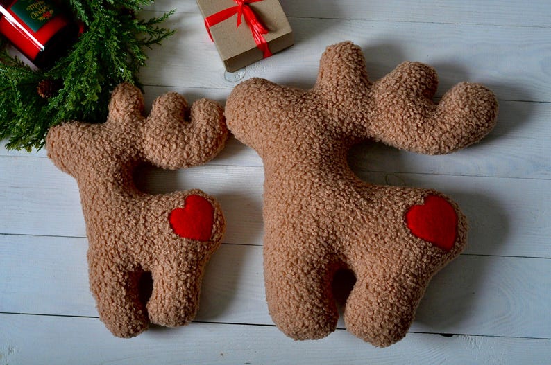 Puede incluir: Dos cojines de peluche marrones con forma de reno, hechos de tela sherpa texturizada. Cada reno presenta un parche de coraz&oacute;n de fieltro rojo brillante en el costado. Los cojines tienen astas distintivas y cuatro patas, con un dise&ntilde;o relleno.