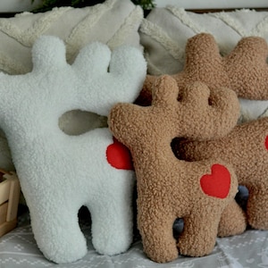 May include: A collection of plush reindeer-shaped pillows made from textured white and tan sherpa fabric. Each reindeer features a bright red heart-shaped patch on its side. The pillows have distinct antlers and four legs, showcasing a soft, fuzzy surface texture throughout the design.