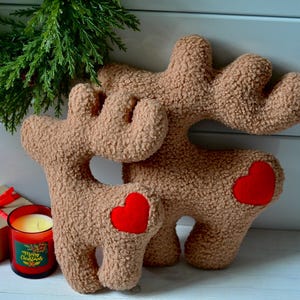 Puede incluir: Dos figuras de reno de peluche en color tostado con textura tipo sherpa, cada una con un parche de coraz&oacute;n rojo brillante en el costado. Los renos tienen astas prominentes y est&aacute;n colocados junto a una peque&ntilde;a caja de regalo con un lazo rojo y una vela roja.