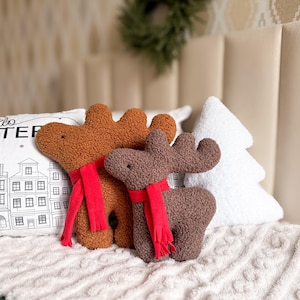May include: Two plush reindeer toys made of textured brown and dark brown fabric, each wearing a bright red scarf. The reindeer have simple embroidered eyes and prominent antlers. They are positioned in front of a white pillow with a black line drawing of a winter cityscape and the text "hello WINTER", alongside a white tree-shaped pillow.