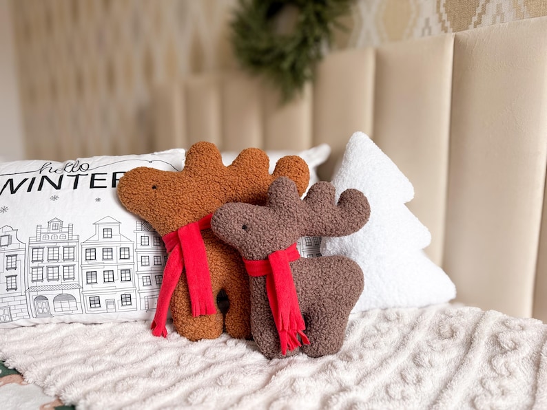 May include: Two plush reindeer toys made of textured brown and dark brown fabric, each wearing a bright red scarf. The reindeer have simple embroidered eyes and prominent antlers. They are positioned in front of a white pillow with a black line drawing of a winter cityscape and the text "hello WINTER", alongside a white tree-shaped pillow.