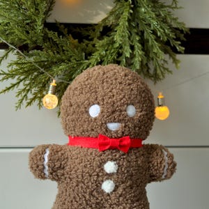 May include: A brown gingerbread man plush toy with a red bow tie and white buttons. The toy is sitting on a white fluffy surface in front of a green Christmas wreath with lights.