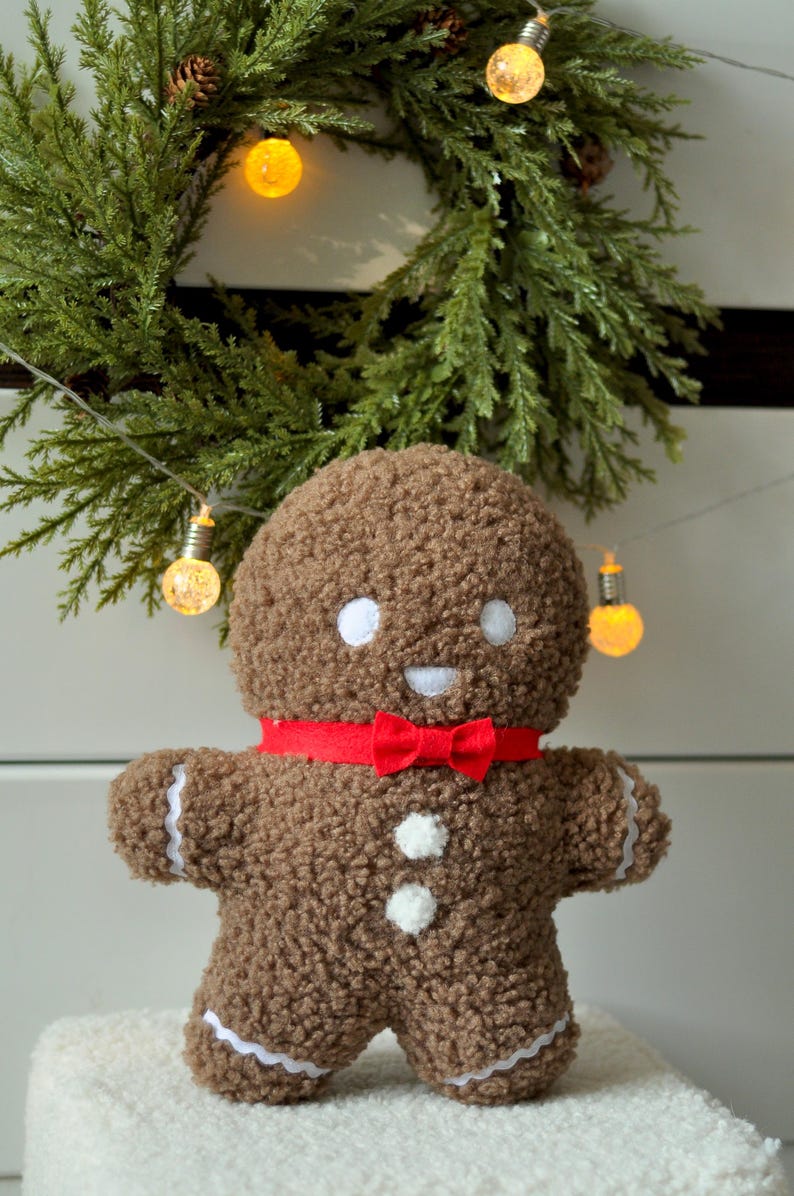 May include: A brown gingerbread man plush toy with a red bow tie and white buttons. The toy is sitting on a white fluffy surface in front of a green Christmas wreath with lights.