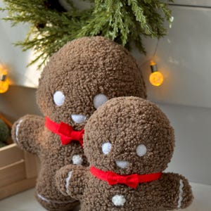 May include: Two brown gingerbread man plush toys with red bow ties. The larger gingerbread man is standing behind the smaller gingerbread man.