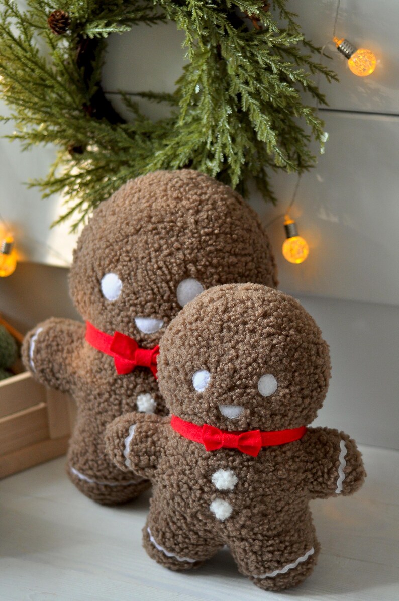May include: Two brown gingerbread man plush toys with red bow ties. The larger gingerbread man is standing behind the smaller gingerbread man.