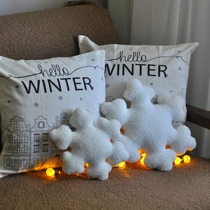 May include: Two white throw pillows with the text "hello winter" on them. The pillows are on a brown couch with white fluffy snowflake-shaped pillows in front of them.