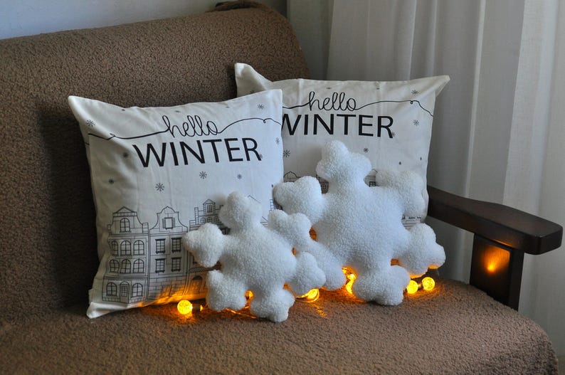 May include: Two white throw pillows with the text "hello winter" on them. The pillows are on a brown couch with white fluffy snowflake-shaped pillows in front of them.