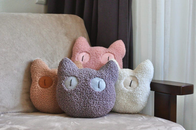 May include: A collection of four cat-shaped throw pillows made of textured, fuzzy sherpa fabric. The pillows are colored in shades of dusty pink, light brown, muted purple, and cream, each featuring two embroidered eyes with vertical black pupils.