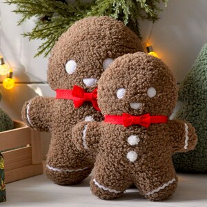 May include: Two gingerbread man plush toys with red bows and white buttons. The larger gingerbread man is standing behind the smaller gingerbread man. Both are sitting on a wooden surface with a green plush Christmas tree behind them.