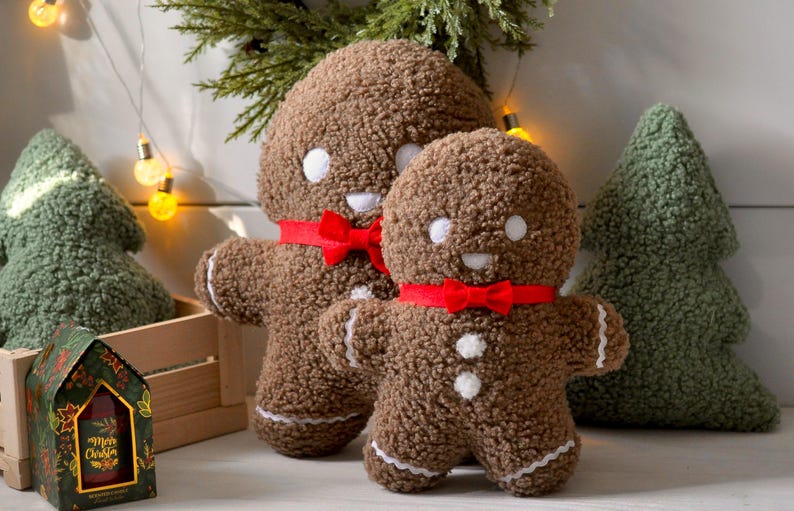 May include: Two gingerbread man plush toys with red bows and white buttons. The larger gingerbread man is standing behind the smaller gingerbread man. Both are sitting on a wooden surface with a green plush Christmas tree behind them.
