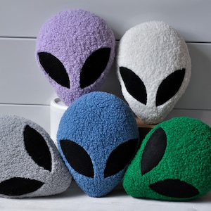 May include: Five plush alien head cushions in shades of purple, white, blue, grey, and green. Each cushion has large black oval eyes. The cushions are made of a soft, textured fabric.