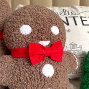 May include: A brown textured plush gingerbread person figure featuring two white circular eyes, a white mouth, two white circular buttons on the torso, and a bright red felt bow tie around the neck. The figure has a soft, fuzzy, sherpa-like fabric.