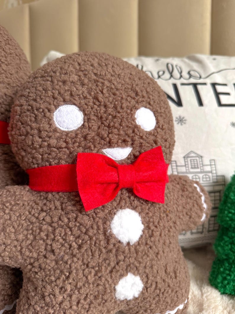May include: A brown textured plush gingerbread person figure featuring two white circular eyes, a white mouth, two white circular buttons on the torso, and a bright red felt bow tie around the neck. The figure has a soft, fuzzy, sherpa-like fabric.