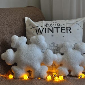May include: Two white plush snowflake-shaped pillows with the text "hello winter" on a white pillow behind them. The pillows are on a brown couch with string lights.