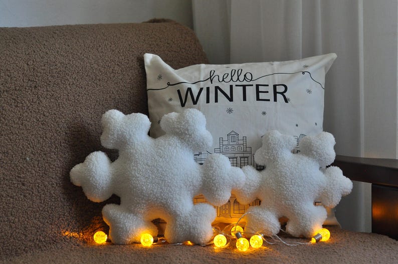 May include: Two white plush snowflake-shaped pillows with the text "hello winter" on a white pillow behind them. The pillows are on a brown couch with string lights.