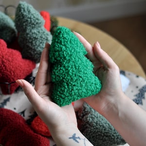 Puede incluir: Un adorno de árbol de Navidad verde y afelpado sostenido en las manos. Otros adornos de árbol de Navidad de felpa en rojo, verde y gris están en una cesta de alambre. Los adornos están sobre una mesa con un camino de mesa festivo.