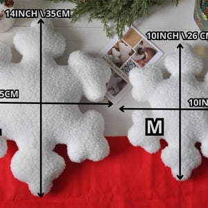 May include: Two white plush snowflake pillows, one large and one medium. The large pillow measures 14 inches by 14 inches, and the medium pillow measures 10 inches by 10 inches. Both pillows are made of soft, fluffy material.