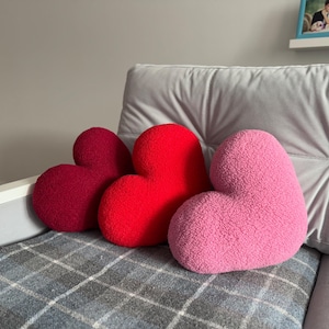 May include: Three heart-shaped cushions in shades of pink, red, and burgundy. The cushions are arranged on a grey sofa with a plaid throw blanket.