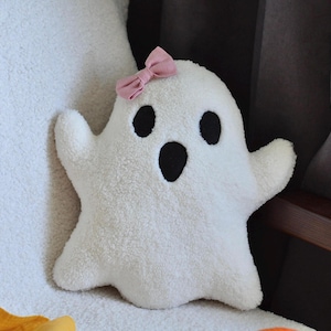 May include: A white ghost-shaped plush pillow made of textured sherpa fabric. The ghost features two black oval eyes and a black oval mouth. A small, light pink fabric bow is attached to the top left side of the ghost's head.