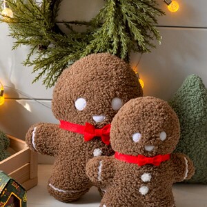 May include: Two plush gingerbread people with red bows, one larger than the other, sitting on a wooden surface. The gingerbread people are brown with white icing details and a smiling face.