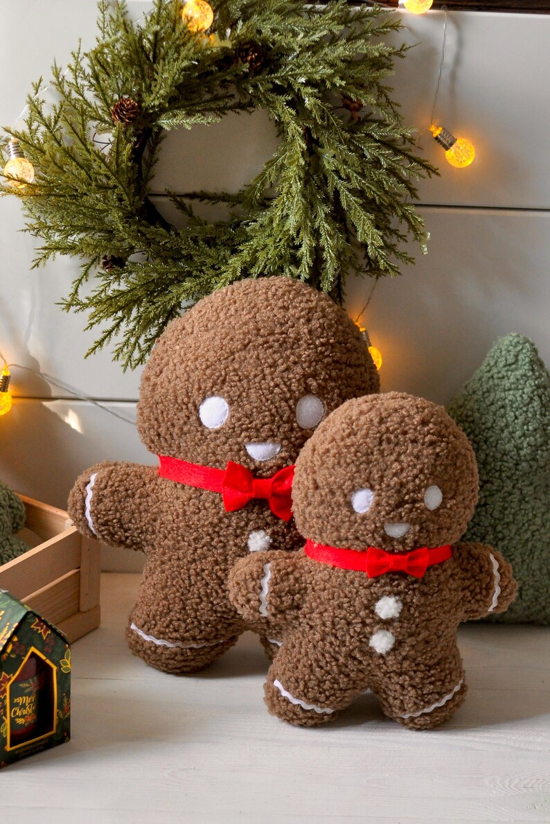 May include: Two plush gingerbread people with red bows, one larger than the other, sitting on a wooden surface. The gingerbread people are brown with white icing details and a smiling face.