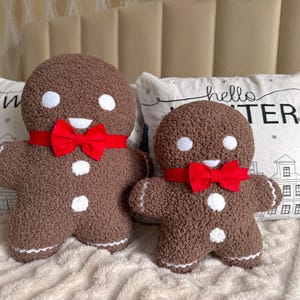 May include: Two brown gingerbread man-shaped cushions with white accents and red bow ties. The cushions have a textured appearance and are placed on a bed with decorative cushions in the background. One cushion is larger than the other.