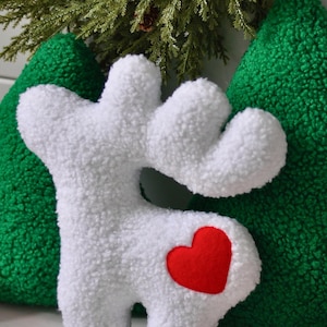 Puede incluir: Un coj&iacute;n de peluche blanco con forma de reno y una superficie texturizada similar al vell&oacute;n rizado. El reno presenta un parche de fieltro rojo brillante en forma de coraz&oacute;n en el costado. Detr&aacute;s hay dos cojines verdes texturizados.