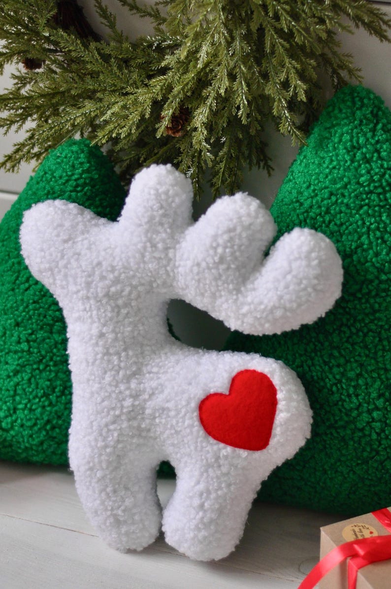 Puede incluir: Un coj&iacute;n de peluche blanco con forma de reno y una superficie texturizada similar al vell&oacute;n rizado. El reno presenta un parche de fieltro rojo brillante en forma de coraz&oacute;n en el costado. Detr&aacute;s hay dos cojines verdes texturizados.