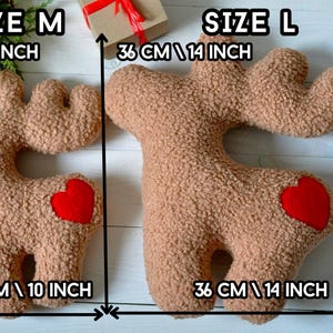 Puede incluir: Dos cojines con forma de reno en tela sherpa texturizada de color canela con un parche de coraz&oacute;n rojo en la cadera. El coj&iacute;n mediano mide 27 cm y el grande mide 36 cm. Las etiquetas de texto indican los tama&ntilde;os SIZE M y SIZE L.