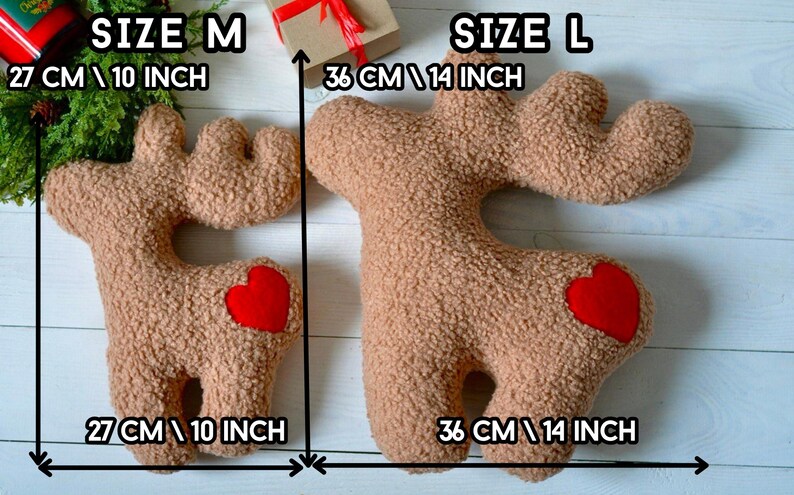 Puede incluir: Dos cojines con forma de reno en tela sherpa texturizada de color canela con un parche de coraz&oacute;n rojo en la cadera. El coj&iacute;n mediano mide 27 cm y el grande mide 36 cm. Las etiquetas de texto indican los tama&ntilde;os SIZE M y SIZE L.