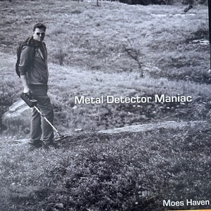 Puede incluir: Imagen en blanco y negro de una persona usando un detector de metales en un campo cubierto de hierba. El texto "Metal Detector Maniac" se muestra en la imagen. La imagen es de Moes Haven.