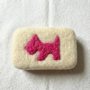 May include: A rectangular bar of soap with a cream-colored base and a bright pink embroidered dog design. The soap is set against a white, textured background, highlighting the handmade quality.