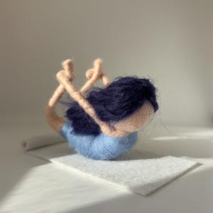 May include: A felted yoga figure in the bow pose, with dark blue hair and a light blue outfit. The figure is on a white felt mat. The arms and legs are a light tan colour.