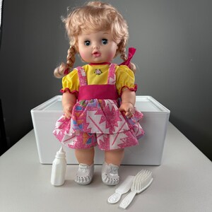 May include: A vintage doll with blonde pigtails, wearing a yellow top, pink overalls, and a patterned skirt. Accessories include a white bottle, comb, and brush. The doll is standing in front of a white box.