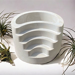 May include: A white concrete planter with five curved shelves for displaying small plants. The planter is sitting on a white surface with two green plants in the background.