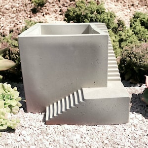 May include: A concrete planter shaped like a building with a set of stairs on the side. The planter is a light gray color and has a rough texture.