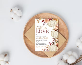 Pode incluir: Um convite de casamento com tema de outono com o texto "Fall in Love". Apresenta abóboras brancas, detalhes florais e está colocado numa bandeja de madeira com um envelope de papel kraft.