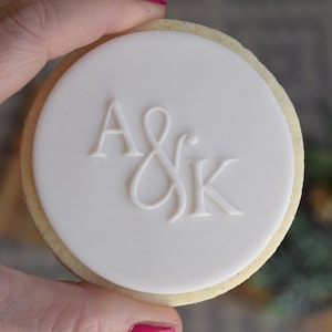 May include: A circular biscuit with a white fondant topping, embossed with the initials "A & K". The biscuit has a light brown edge and is held by a hand. The design suggests a wedding or celebratory theme.