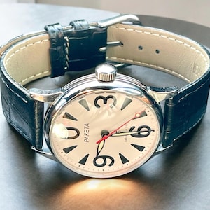 Puede incluir: Un reloj de pulsera plateado con una esfera blanca y números negros. El reloj tiene una correa de cuero negra y una aguja de segundos roja.