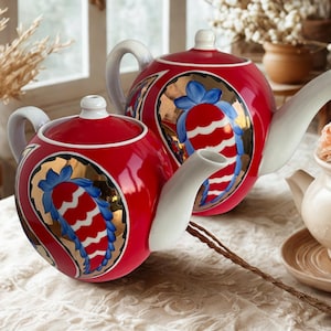 May include: Two red ceramic teapots with white spouts and handles. Each teapot features a gold oval design with a blue bow and a red and white pattern. The teapots are on a white tablecloth.