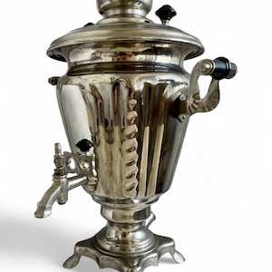 Può includere: Un samovar d'argento vintage con beccuccio decorativo, manici e coperchio. Il samovar ha uno sfiato a forma di stella e poggia su una base a piedistallo. Le maniglie e le manopole sono nere.