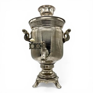 May include: A silver samovar with a decorative spout, handles, and a lid. The tea urn sits on a decorative base with four feet. The samovar is designed to heat water for tea and has a vintage appearance.